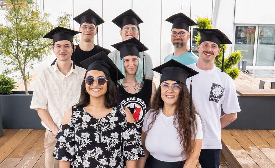Acht junge Erwachsene mit Abschlusskappen stehen lächelnd auf einer Terrasse, im Hintergrund Pflanzen und moderne Gebäude.